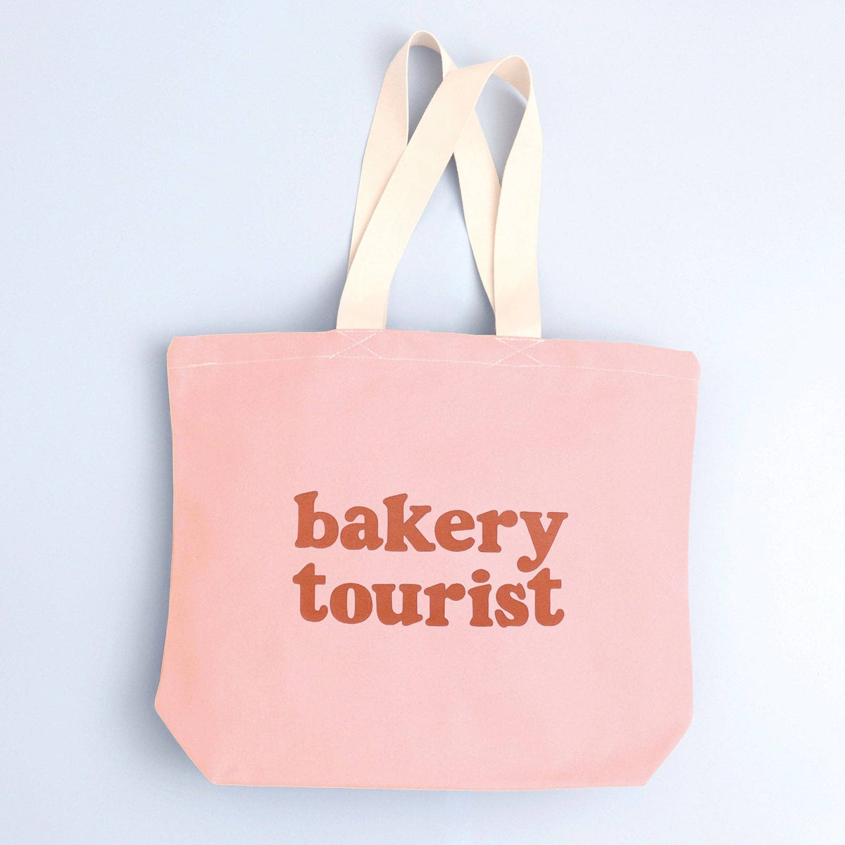Blush Pink Tote Bag with ‘Bakery Tourist ’ text on a Blue Background