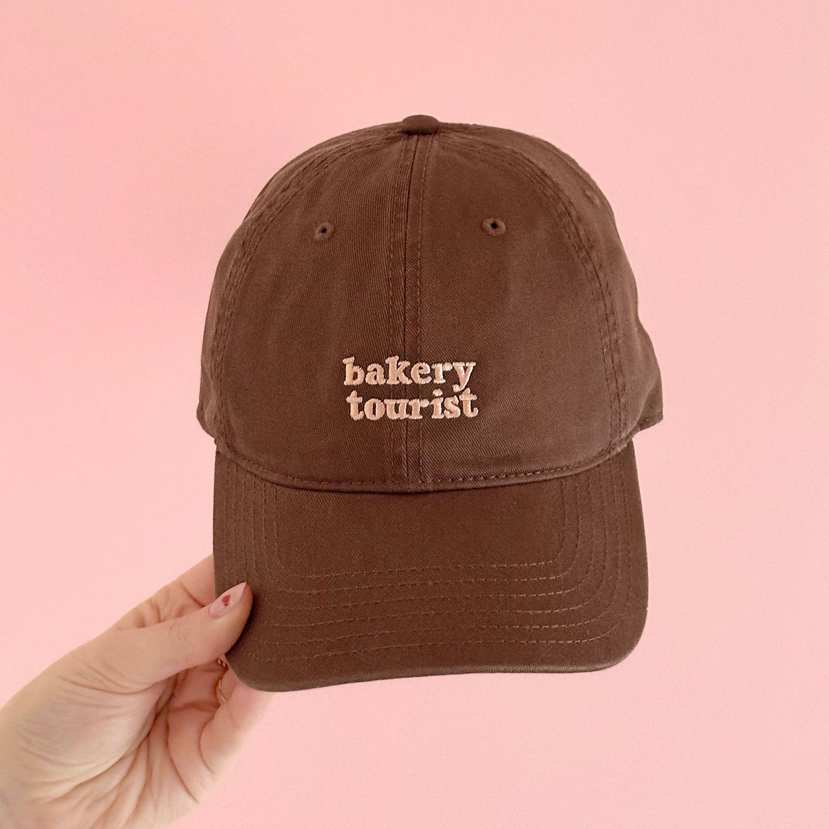 Cocoa Cotton Baseball Cap with ‘Bakery Tourist’ Text on a Pink Background