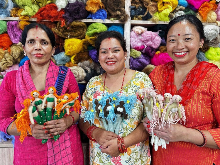 Three women holding handmade dolls in front of a colorful display of yarn and fabric.