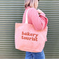 Person Carrying a Blush Pink Tote Bag with ‘Bakery Tourist ’ text on a Green Background