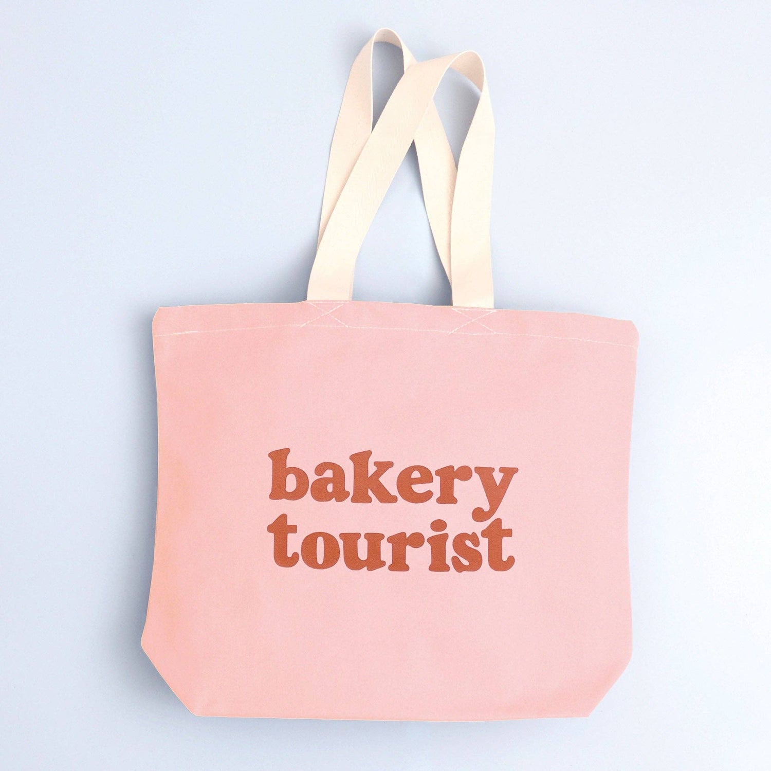 Blush Pink Tote Bag with ‘Bakery Tourist ’ text on a Blue Background