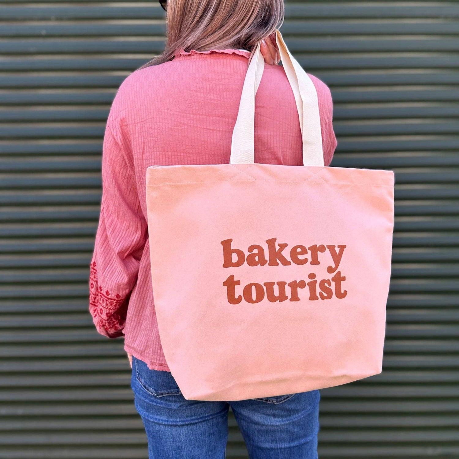 Person Carrying a Blush Pink Tote Bag with ‘Bakery Tourist ’ text on a Green Background