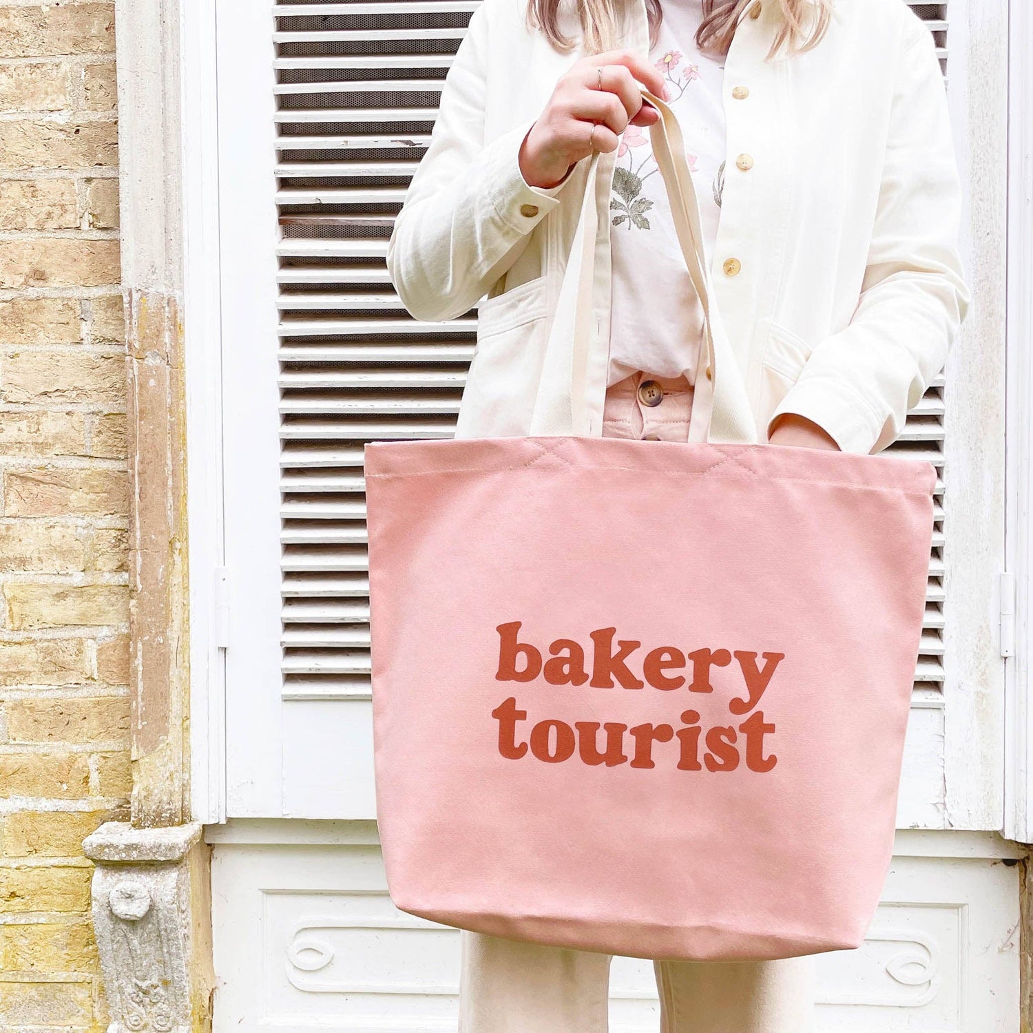 Person Carrying a Blush Pink Tote Bag with ‘Bakery Tourist ’ text on a mixed Background
