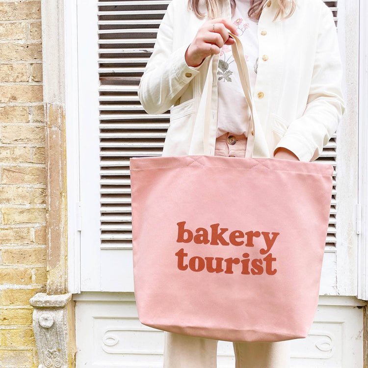 Person Carrying a Blush Pink Tote Bag with ‘Bakery Tourist ’ text on a mixed Background