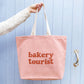 Blush Pink Tote Bag with ‘Bakery Tourist ’ text on a Blue Background
