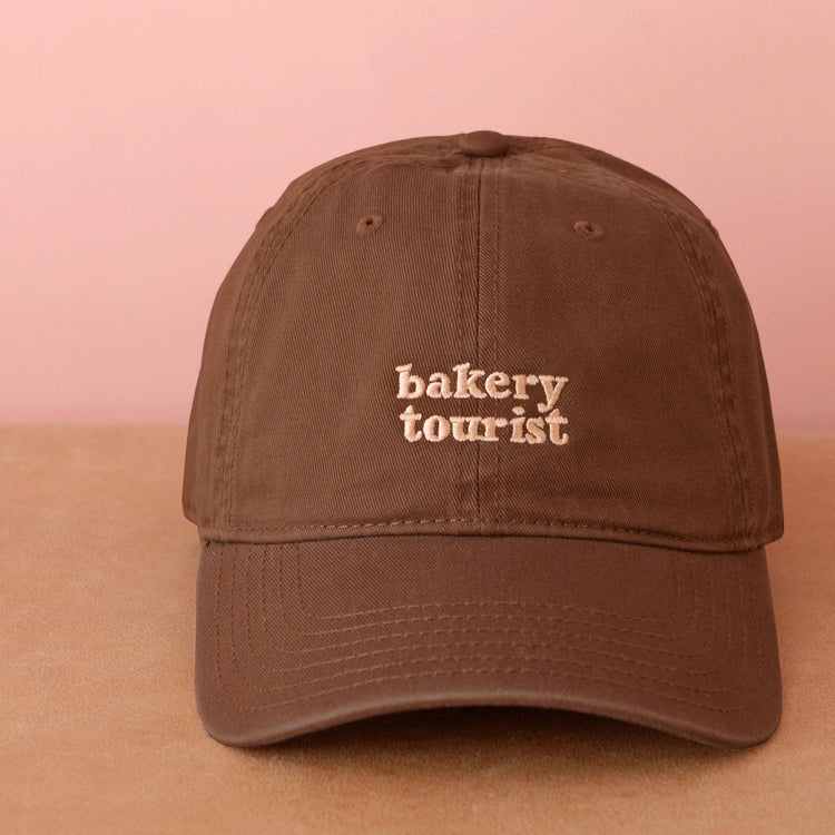 Cocoa Cotton Baseball Cap with ‘Bakery Tourist’ Text on a Neutral Background
