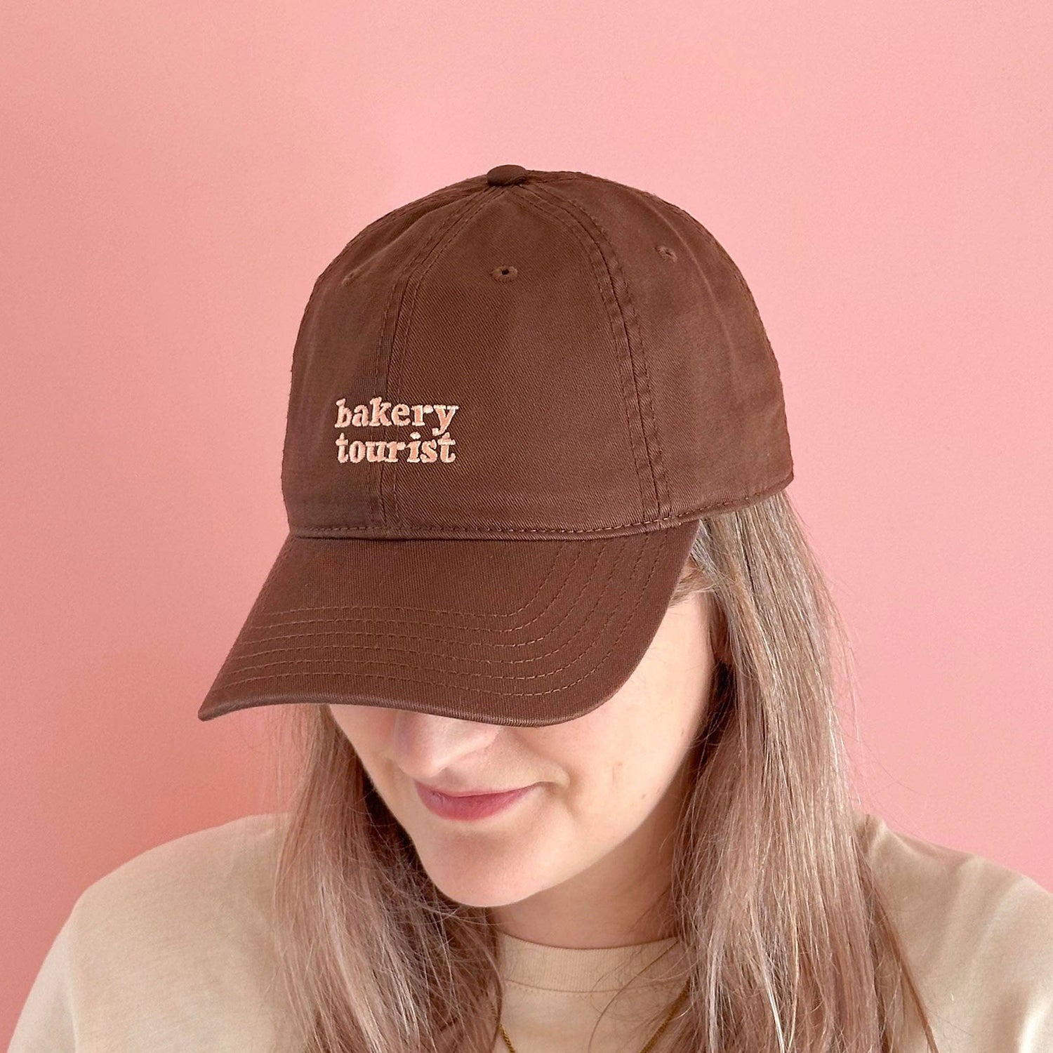 Person Wearing a Cocoa Cotton Baseball Cap with ‘Bakery Tourist’ Text on a Pink Background