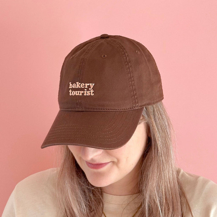 Person Wearing a Cocoa Cotton Baseball Cap with ‘Bakery Tourist’ Text on a Pink Background