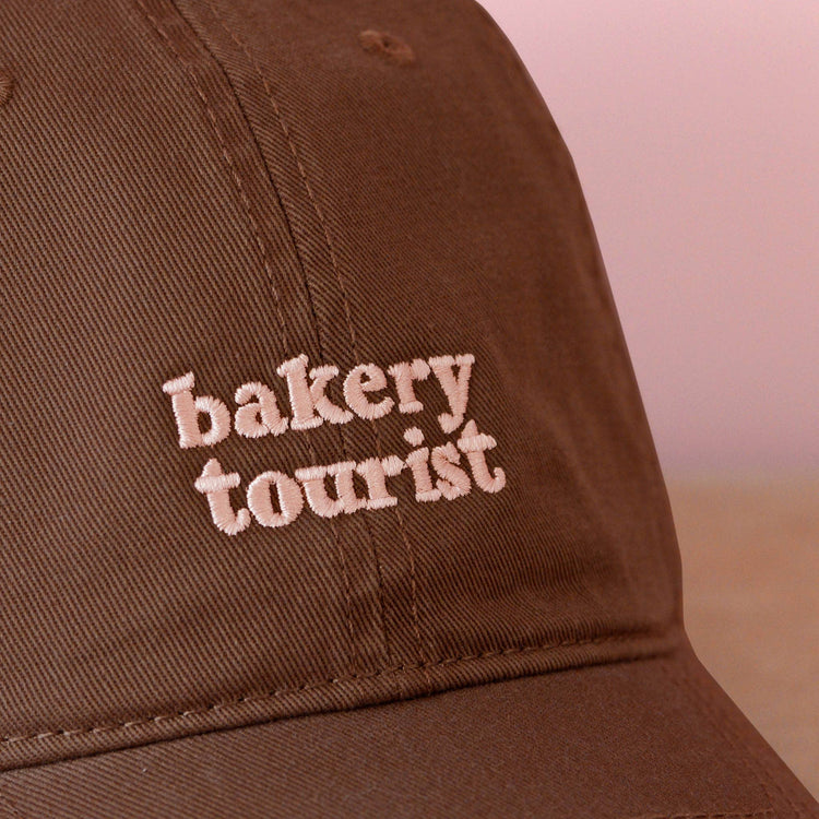 Cocoa Cotton Baseball Cap with ‘Bakery Tourist’ Text on a pink Background