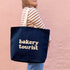 Person Carrying a Midnight Blue Tote Bag with ‘Bakery Tourist’ Text on a pink Background