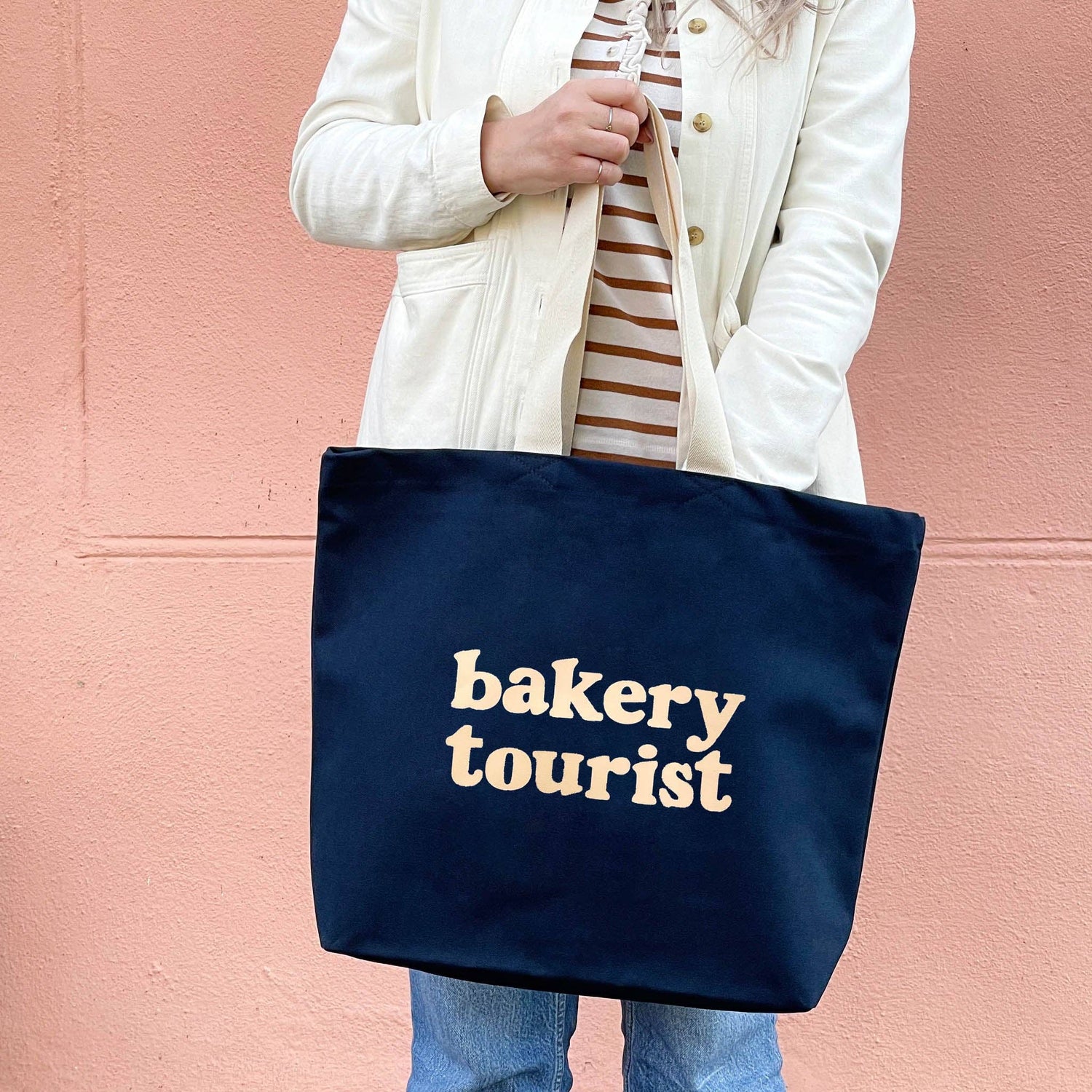 Person Carrying a Midnight Blue Tote Bag with ‘Bakery Tourist’ Text on a Pink Background