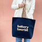 Person Carrying a Midnight Blue Tote Bag with ‘Bakery Tourist’ Text on a Pink Background
