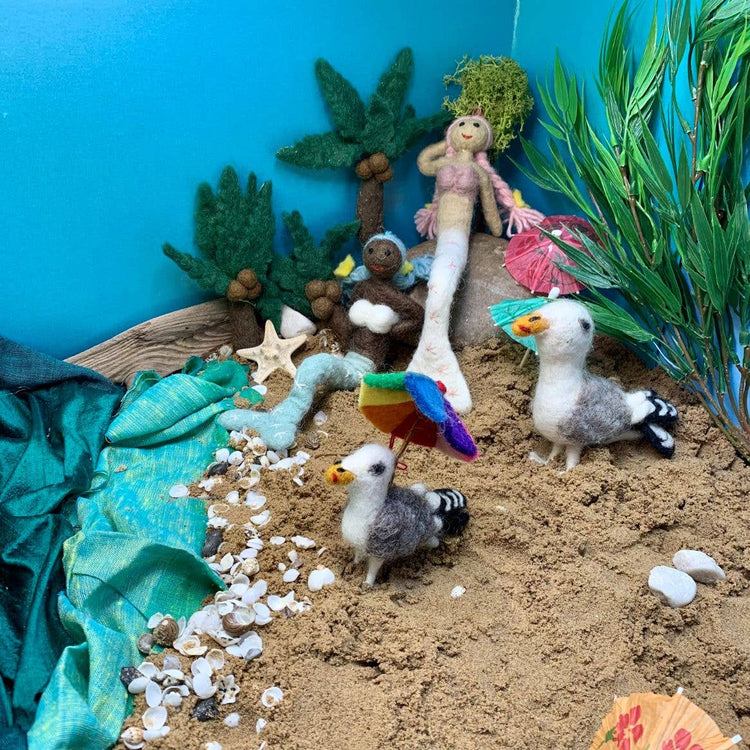 Miniature beach scene with toy mermaids, seagulls, and tropical plants against a blue wall.
