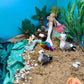 Miniature beach scene with toy mermaids, seagulls, and tropical plants against a blue wall.