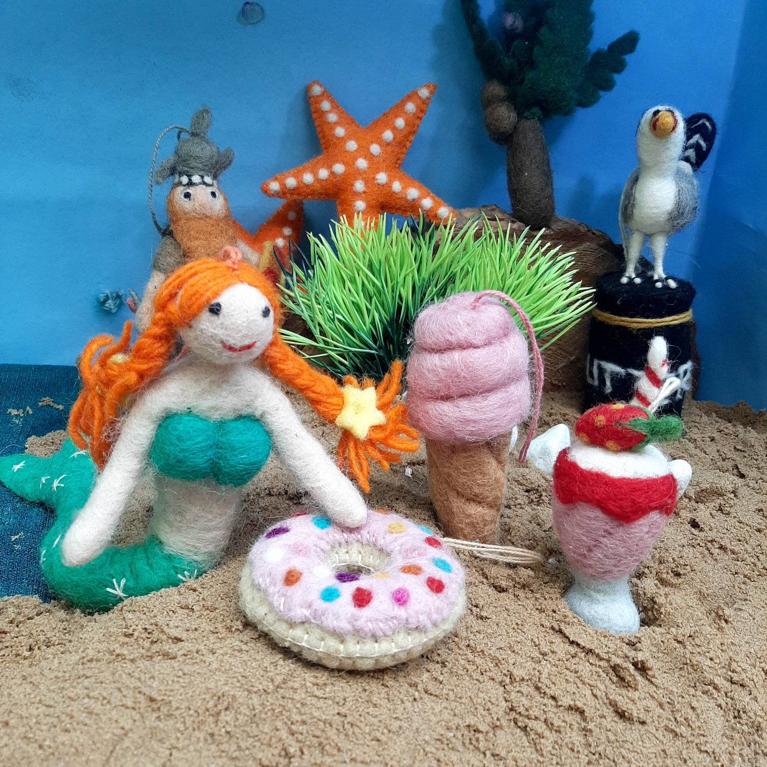 Miniature mermaid figurine with ice cream and donut on a sandy surface with starfish and grass.