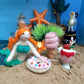 Miniature mermaid figurine with ice cream and donut on a sandy surface with starfish and grass.