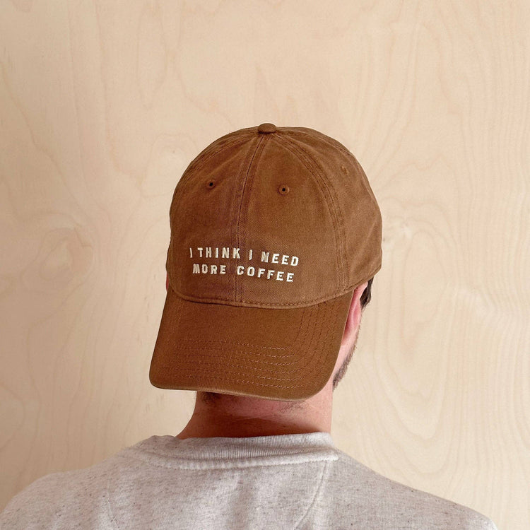 Person Wearing a Cotton Baseball Cap with ‘I Think I Need More Coffee’ Text on a White Background