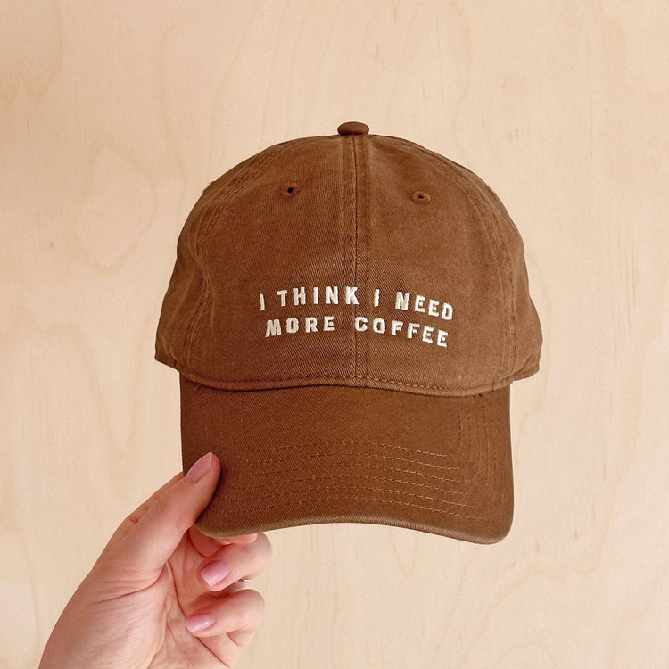 Cotton Baseball Cap with ‘I Think I Need More Coffee’ Text on a Neutral Background
