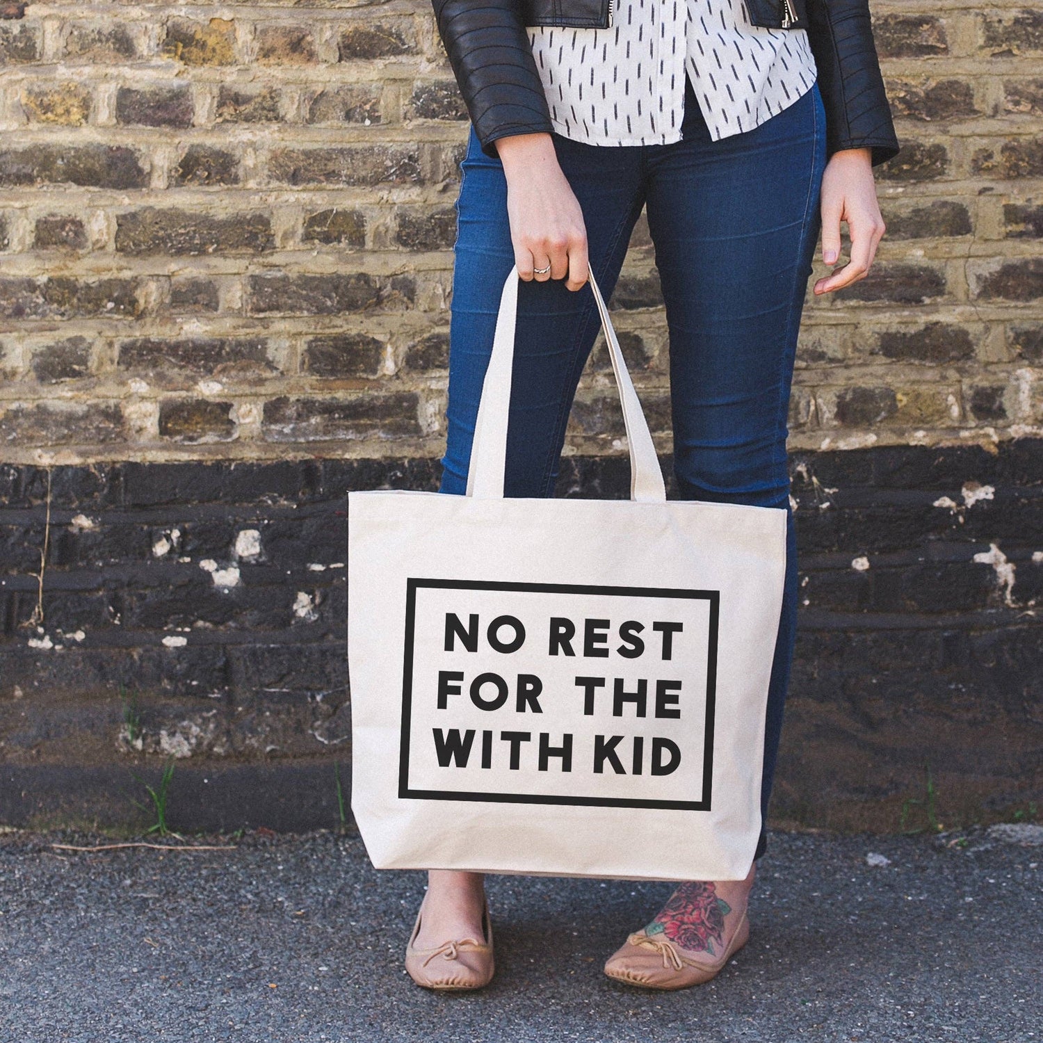 Person Carrying a Canvas Tote Bag with ‘No Rest For With the Kid’ Text on a Neutral Background