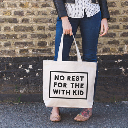 Person Carrying a Canvas Tote Bag with ‘No Rest For With the Kid’ Text on a Neutral Background