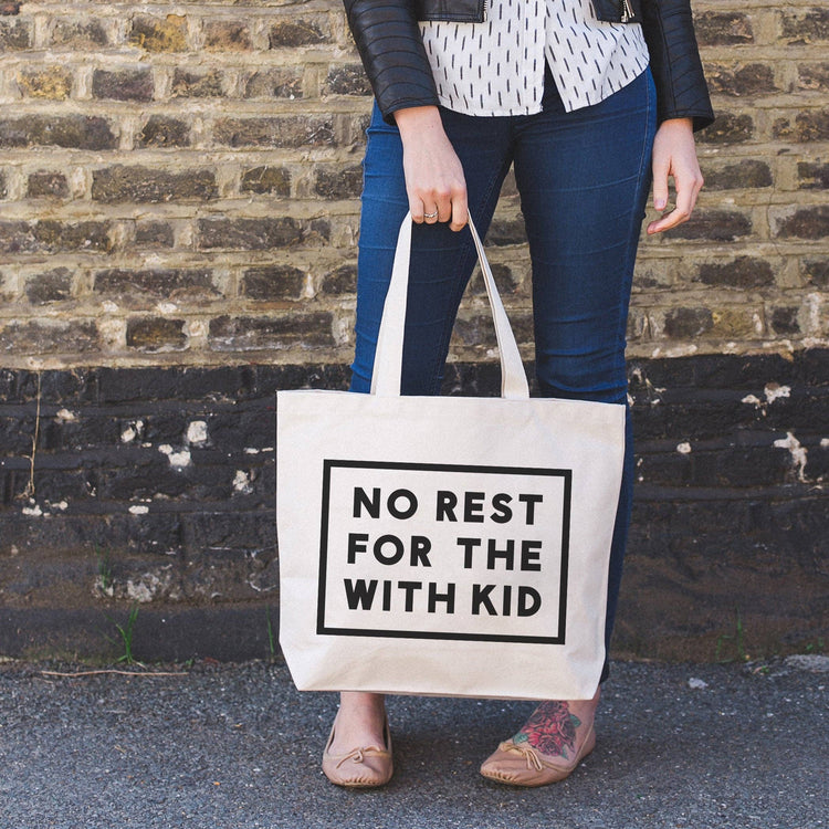 Person Carrying a Canvas Tote Bag with ‘No Rest For With the Kid’ Text on a Neutral Background