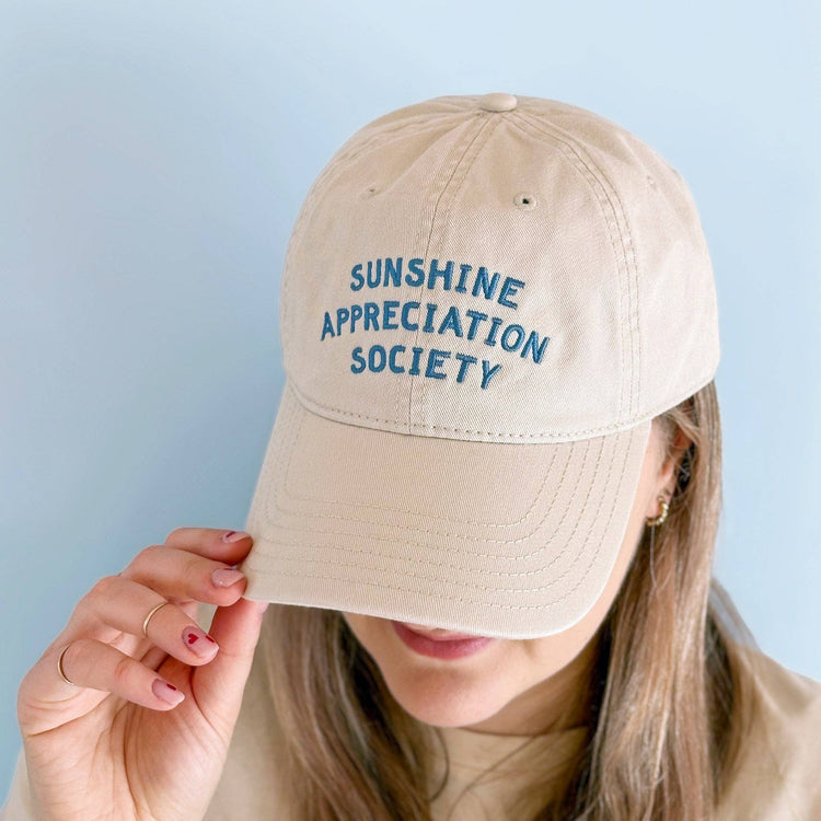 Person Wearing an Off-White Cotton Baseball Cap with ‘Sunshine Appreciation Society’ text on a Blue Background