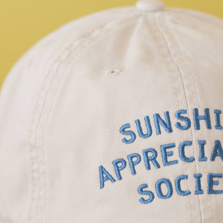 Off-White Cotton Baseball Cap with ‘Sunshine Appreciation Society’ text on a Neutral Background