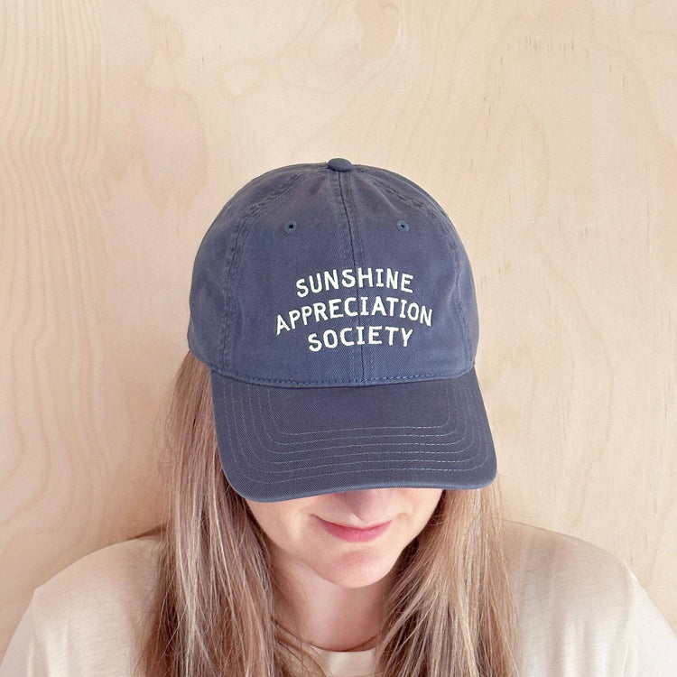 Person Wearing a Vintage Blue Cotton Baseball Cap with ‘Sunshine Appreciation Society’ Text on a Neutral Background