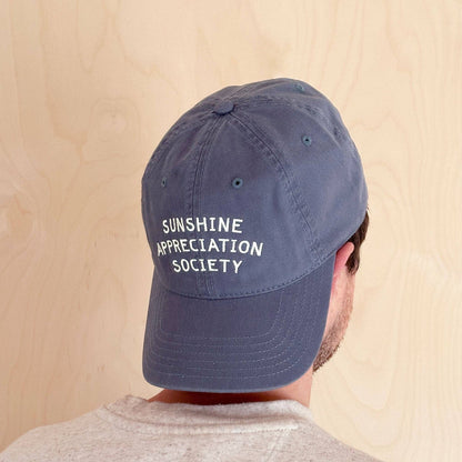 Person Wearing a Vintage Blue Cotton Baseball Cap with ‘Sunshine Appreciation Society’ Text on a Neutral Background