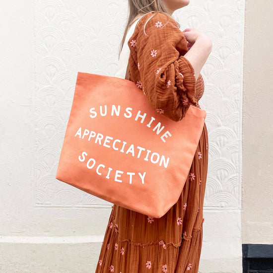 Person Carrying a Peach Canvas Tote Bag with ‘Sunshine Appreciation Society’ text on a Neutral Background