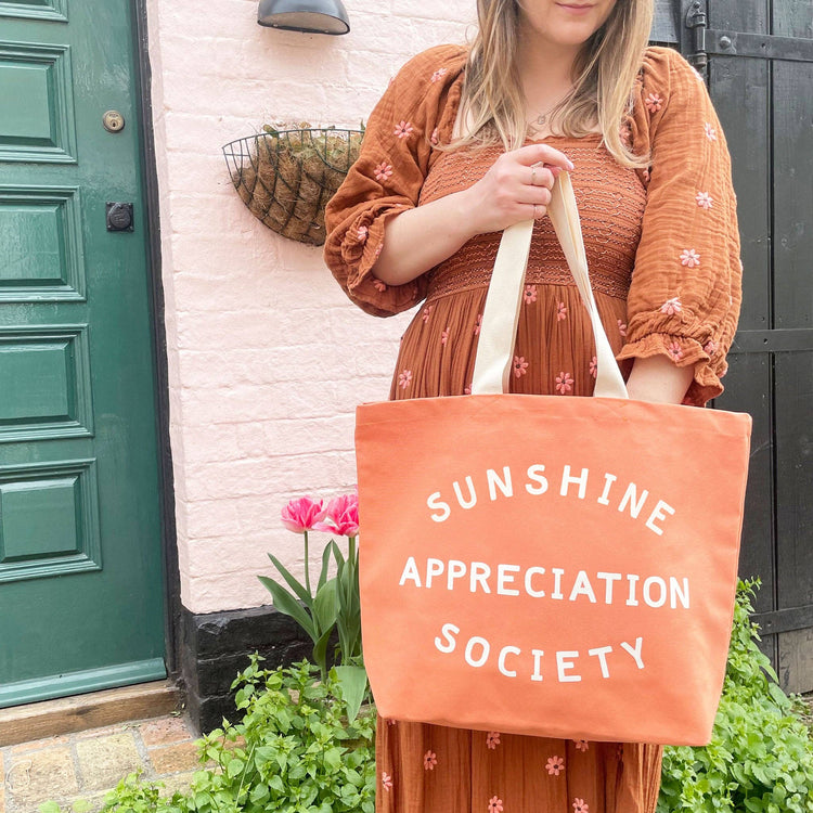 Person Carrying a Peach Canvas Tote Bag with ‘Sunshine Appreciation Society’ text on a Mixed Background