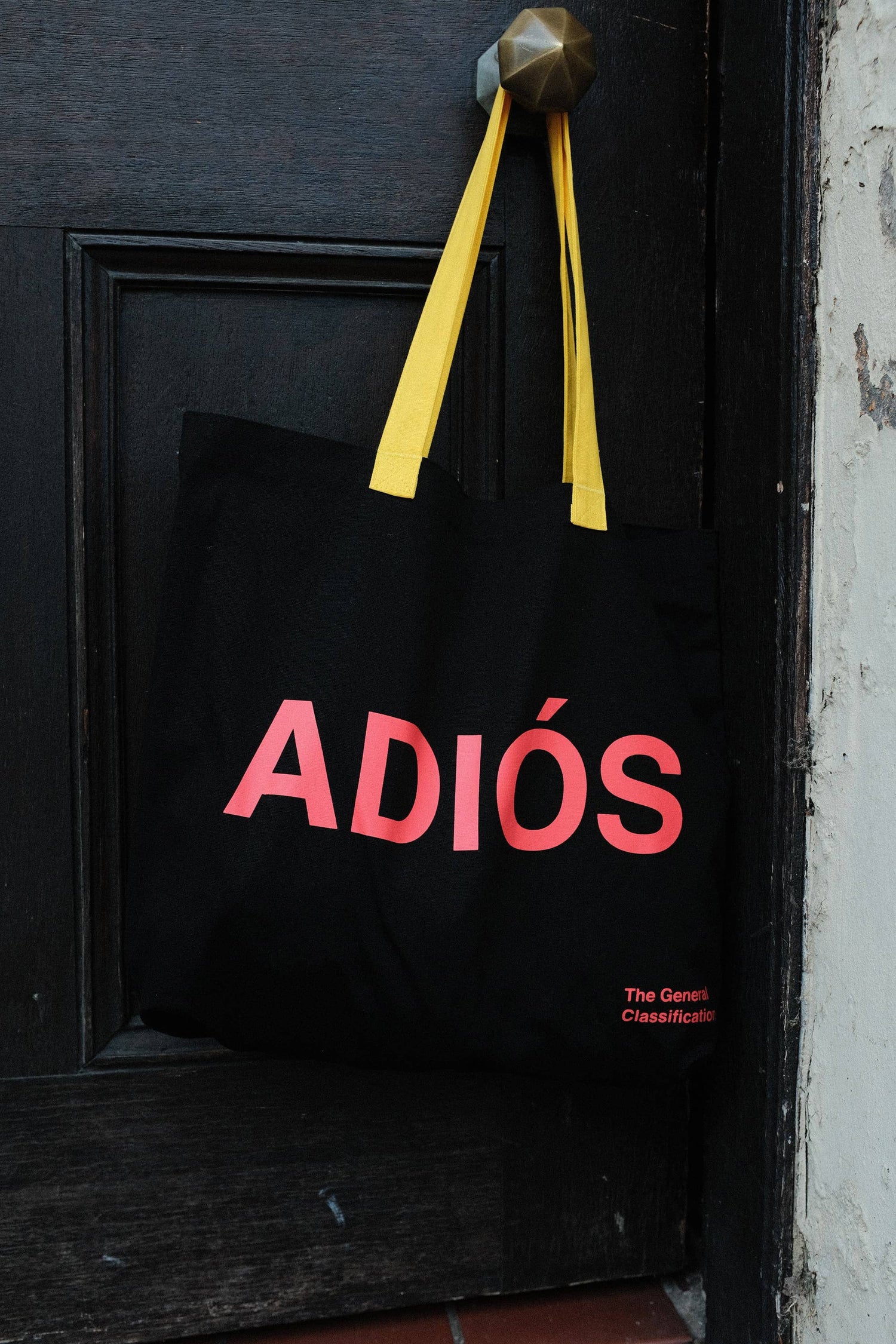 Black reusable canvas tote bag with yellow handles and bold red "Hola" and ‘Adiós’ graphic by The General Classification. Eco-friendly cotton shopping bag with modern minimalist design, ideal for everyday use, groceries, travel, and casual fashion.