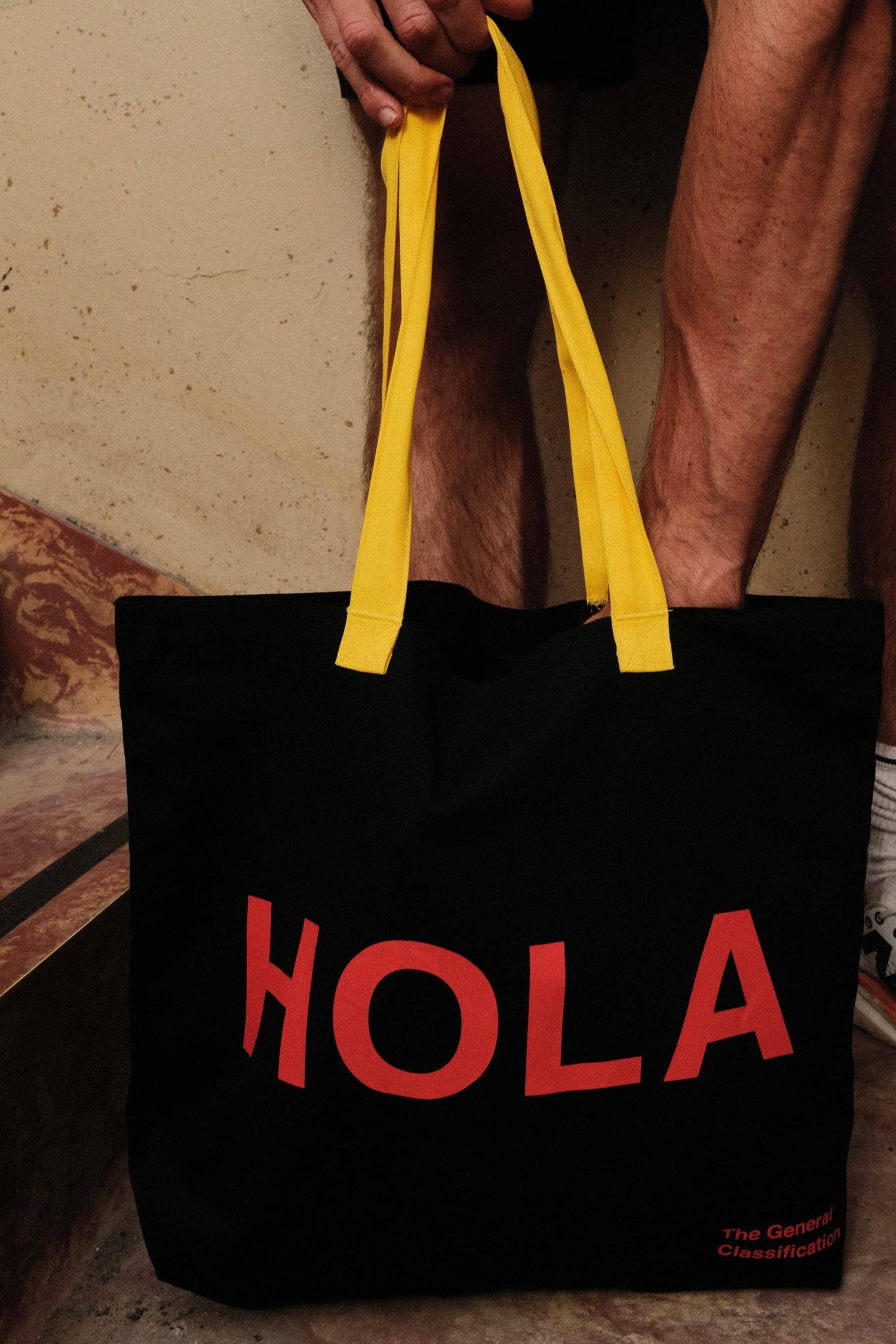 Black reusable canvas tote bag with yellow handles and bold red "Hola" and ‘Adiós’ graphic by The General Classification. Eco-friendly cotton shopping bag with modern minimalist design, ideal for everyday use, groceries, travel, and casual fashion.