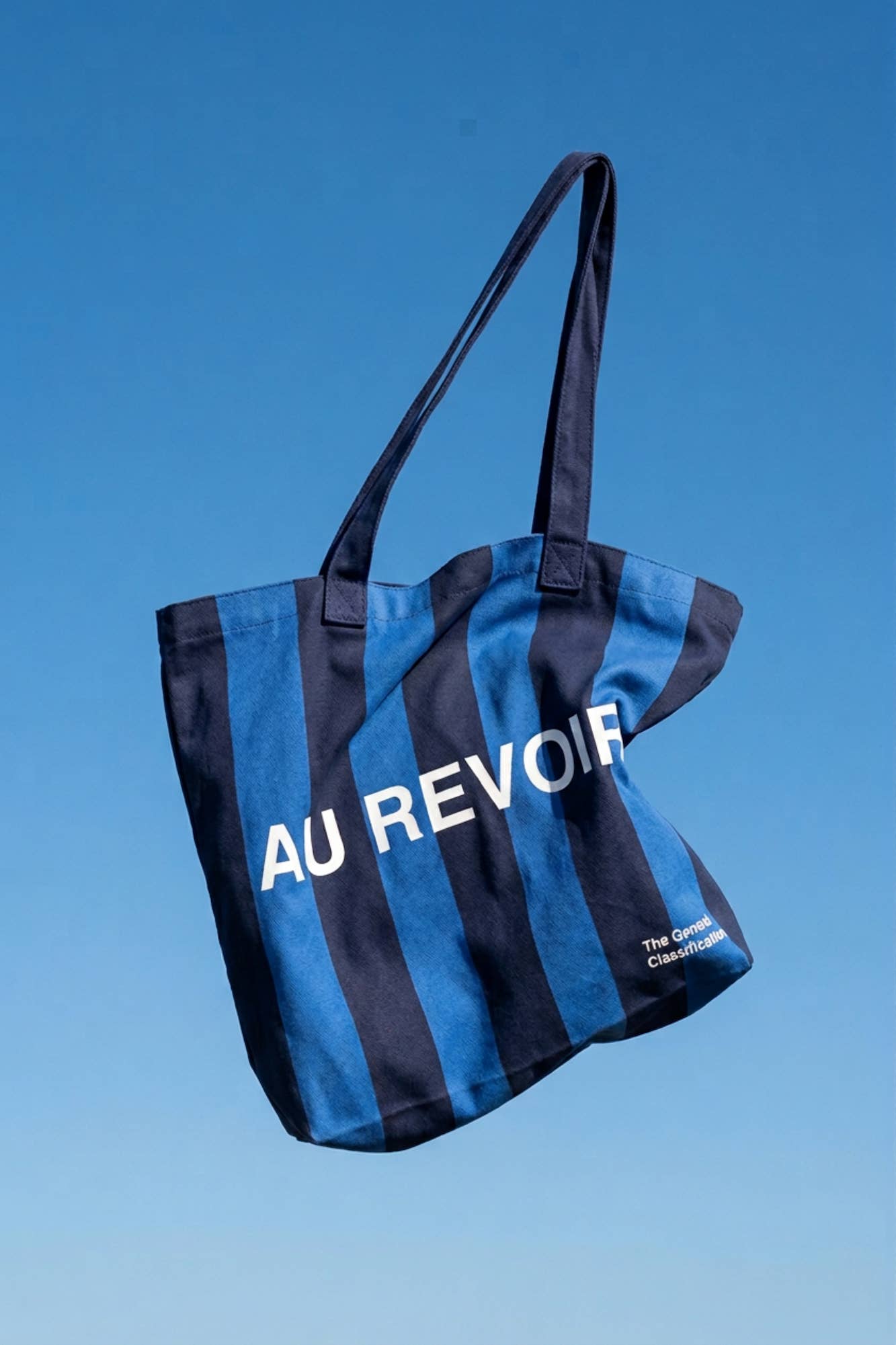 Blue and navy stripe reusable canvas tote bag with navy handles and bold white "Bonjour" and ‘Au Revoir’ graphic by The General Classification. Eco-friendly cotton shopping bag with modern minimalist design, ideal for everyday use, groceries, travel, and casual fashion.
