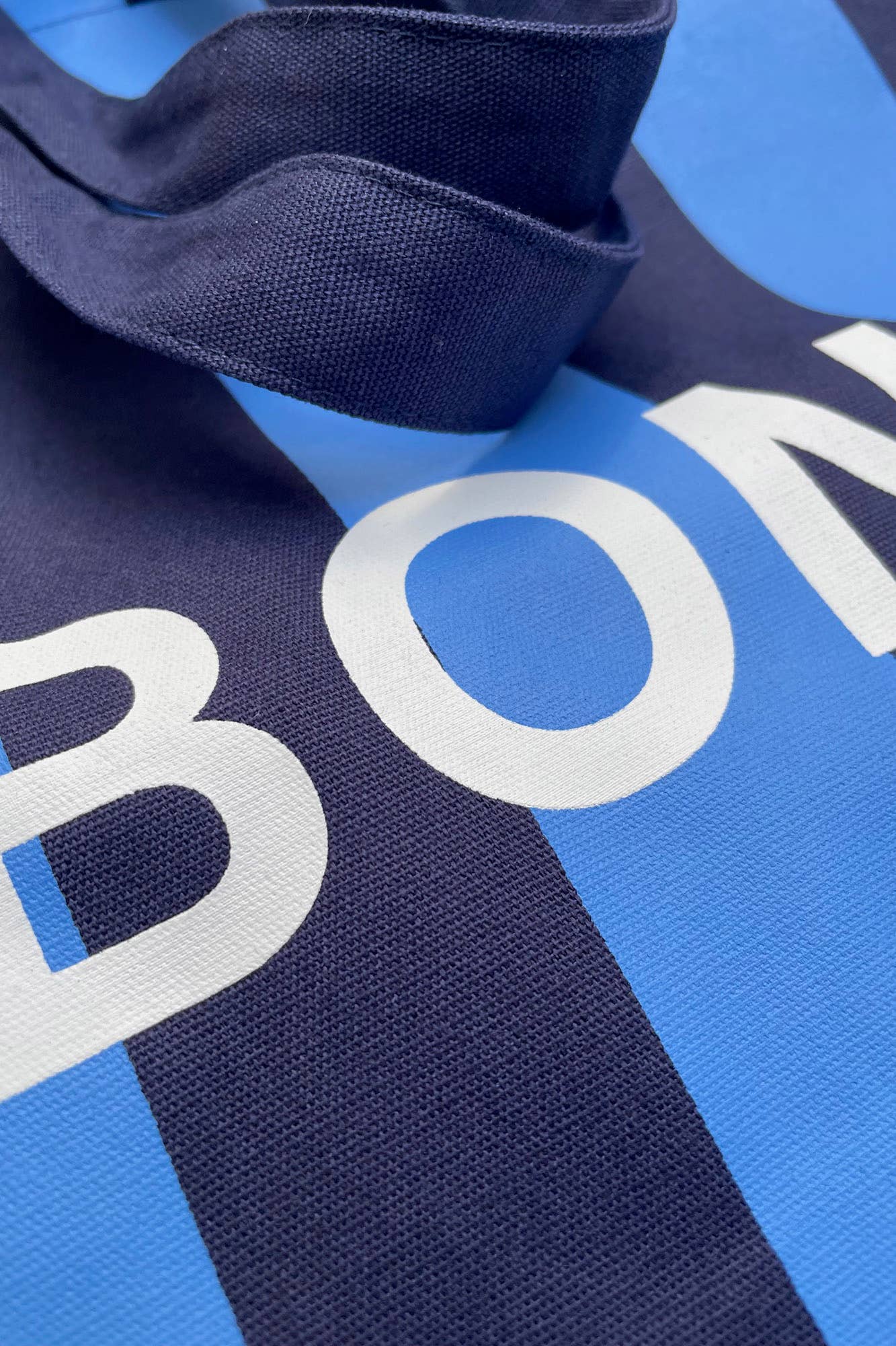 Blue and navy stripe reusable canvas tote bag with navy handles and bold white "Bonjour" and ‘Au Revoir’ graphic by The General Classification. Eco-friendly cotton shopping bag with modern minimalist design, ideal for everyday use, groceries, travel, and casual fashion.